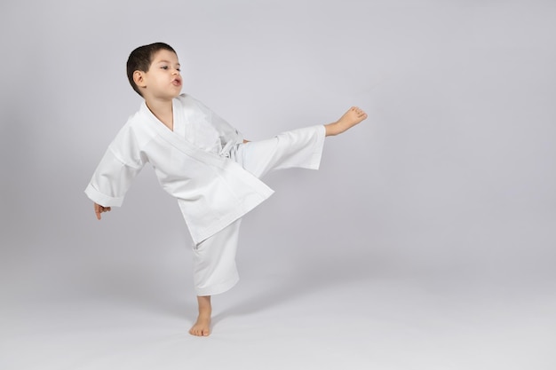 Een kleine jongen in een kimono beoefent karate op een witte achtergrond die naar voren schopt