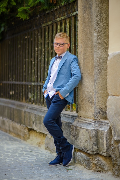 Een kleine jongen in een pak en een bril staat in de buurt van de muur.