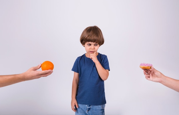 Een kleine jongen in een t-shirt maakt een keuze tussen een sinaasappel en een donut op een witte achtergrond met een plek voor tekst