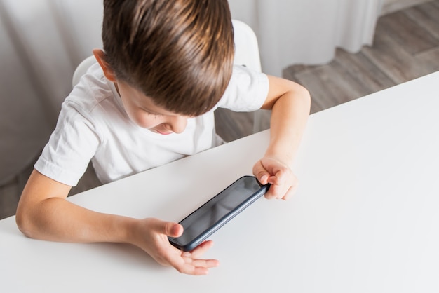 Een kleine jongen in een wit T-shirt speelt thuis spelletjes aan de telefoon. Een gelukkig kind kijkt naar zijn smartphone.