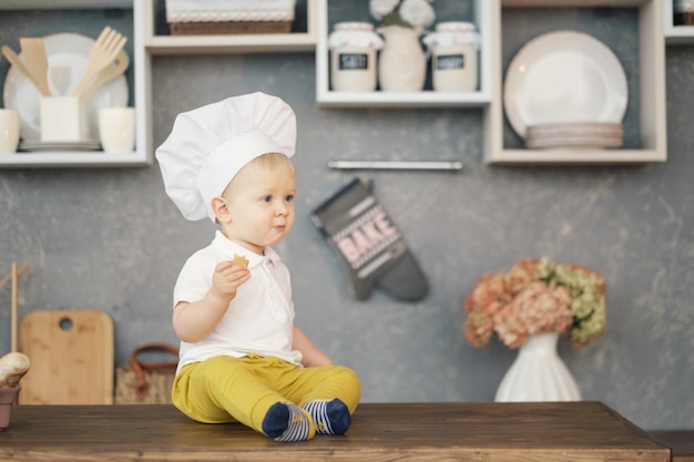 een kleine jongen in hoed van chef-kok in de keuken op de tafel zitten en eten