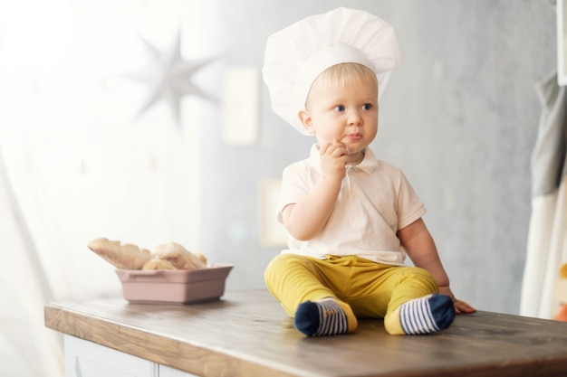 een kleine jongen in hoed van chef-kok in de keuken op de tafel zitten en eten