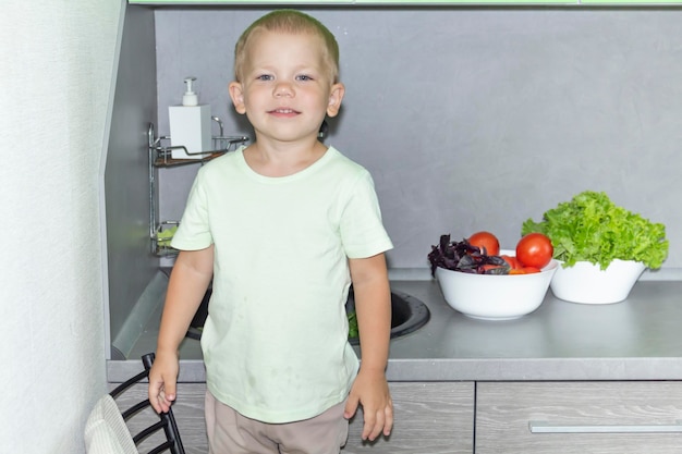 Een kleine jongen met een kort kapsel helpt bij het koken in de keuken Wast verse groenten en kruiden in een zwarte gootsteen in een grijze keuken