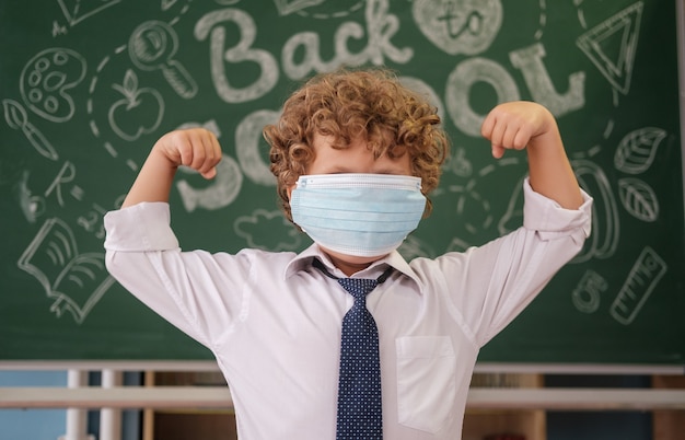 Een kleine jongen met een medisch masker op de achtergrond van een schoolbord met de tekst terug naar school