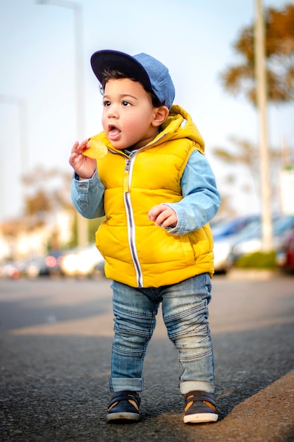 Een kleine jongen op straat in winterkleren en een hoed op zijn hoofd en een stuk chips in zijn hand