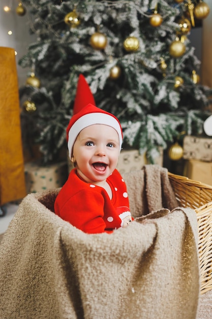 Een kleine jongen van 1 jaar oud zit in een mand tegen de achtergrond van een kerstboom Een kind in een rode nieuwjaarswind bij de kerstboom
