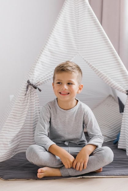 Een kleine jongen zit halverwege in een lichte kinderkamer bij de wigwam en lacht