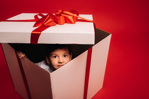 Een kleine jongen zit in een kerstcadeau en kijkt eruit op een rode achtergrond