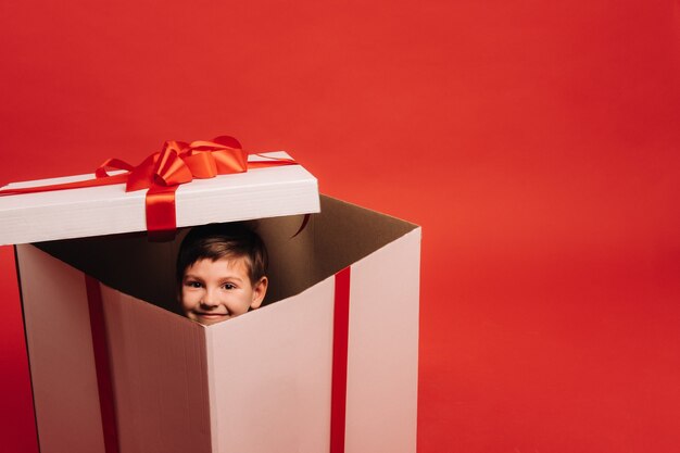 Een kleine jongen zit in een kerstcadeau en kijkt eruit op een rode achtergrond