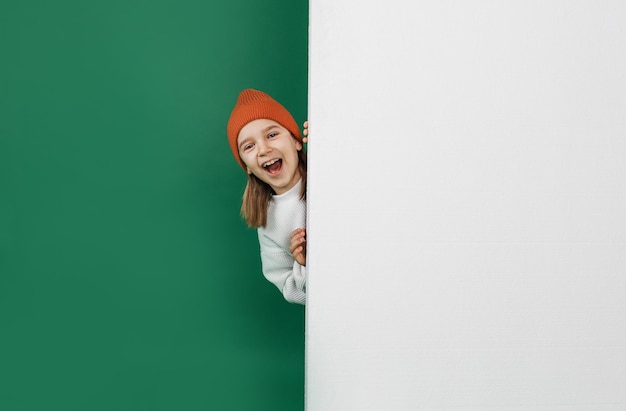 Een kleine knappe jongen, glurend achter een witte muur met een lege ruimte, op een afgelegen groene achtergrond