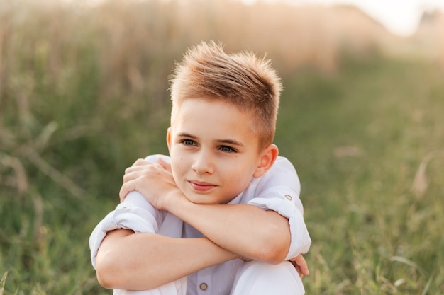 Een kleine knappe jongensblonde in een wit overhemd en een witte broek in de natuur in de zomer. Gelukkige jeugd. Positieve emoties