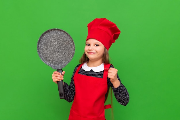 Een kleine meisjeschef-kok met een rode hoed en een schort houdt een pannenkoekenpan vast op een groene geïsoleerde achtergrond Een beroep kiezen voor een student