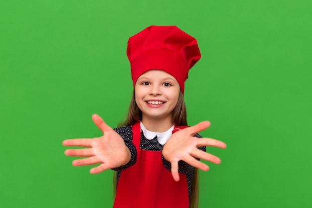 Een kleine meisjeschef-kok strekt haar handen uit met de handpalmen naar voren Een kind gekleed in een rode schort en een kookpet op een groene geïsoleerde achtergrond