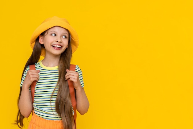 Een kleine meisjesscout in een zomerhoed en met een rugzak kijkt naar uw advertentie Het kind gaat op kampeertrip Gele geïsoleerde lichte achtergrond Kopieer ruimte Banner