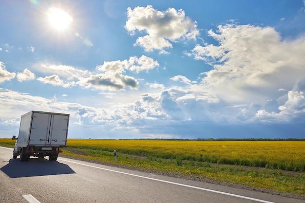 Een kleine vrachtwagen beweegt langs de weg tegen de achtergrond van een blauwe lucht en een geel veld als rug