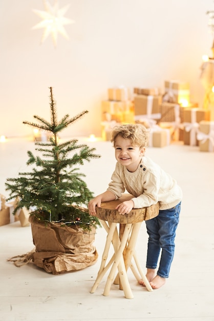Een knap krullend jongetje zit op een houten stoel in de buurt van een kerstboom