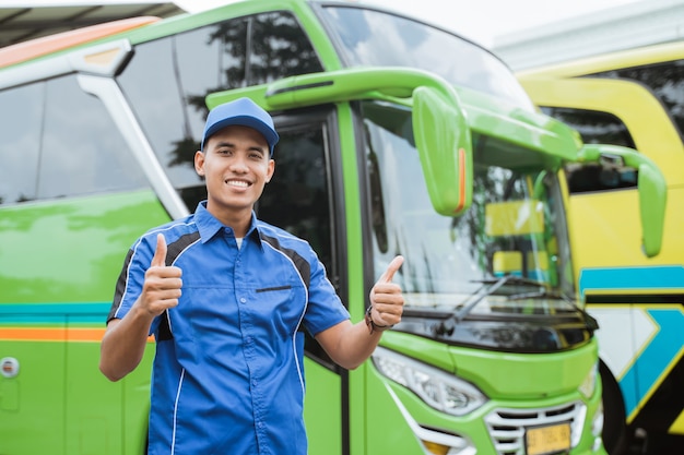 Een knappe buschauffeur in uniform en hoed glimlacht met duimen omhoog tegen de achtergrond van de bus