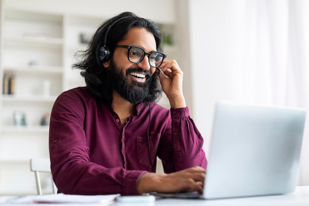 Een knappe jonge Indiase man met een headset en een bril kijkt naar een webinar op een laptop