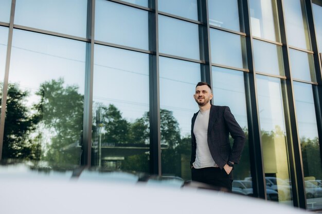 Een knappe jonge zakenman in pak staat bij zijn sportwagen voor het moderne zakencentrum.