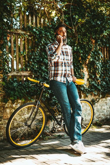 Een knappe jongeman gaat met zijn fiets naar de stad, staat ernaast, wacht op iemand en praat op de smartphone.