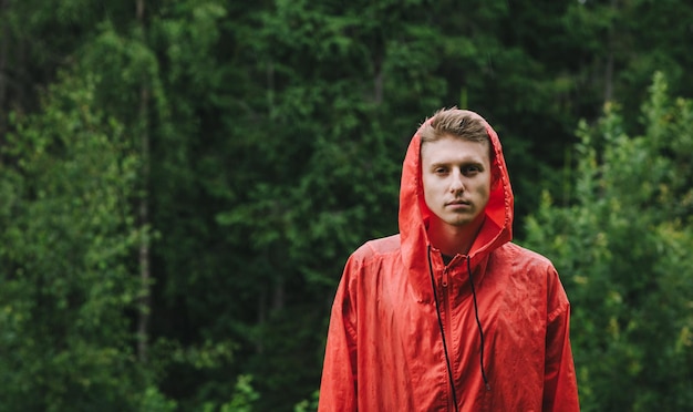 Een knappe jongeman in een rode regenjas staat op de achtergrond van het bos in de regen