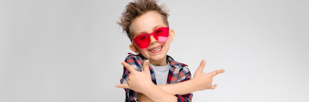 Een knappe jongen in een geruit hemd, grijs shirt en spijkerbroek staat. Een jongen in rode zonnebril. De jongen toont een rocker geit.