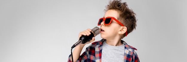 Een knappe jongen in een plaid shirt, grijs shirt en spijkerbroek staat op een grijze achtergrond. Een jongen die een zonnebril draagt. Roodharige jongen zingt in de microfoon