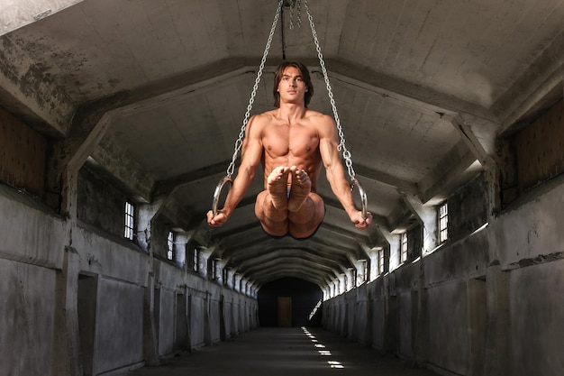 Een knappe professionele turnster met een mooi gespierd lichaam traint op gymnastiekringen in het verlaten industriële gebouw, toont de statische positie