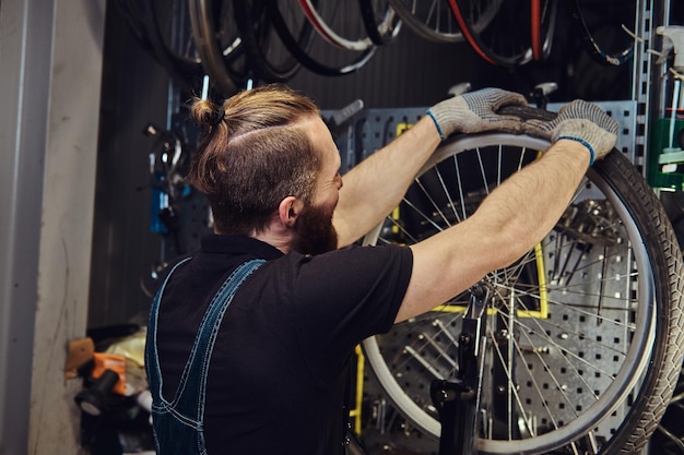 Een knappe roodharige man in een jeans overall, werkend met een fietswiel in een reparatiewerkplaats. een werknemer verwijdert de fietsband in een werkplaats.