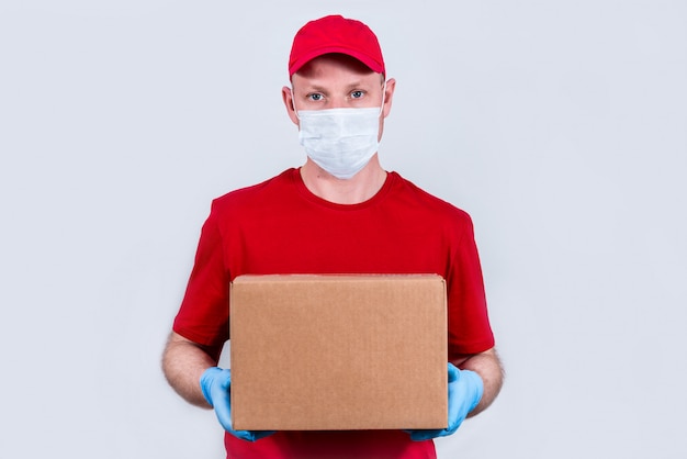 Foto een koerier in een rood uniform en een beschermend medisch masker en handschoenen heeft een kartonnen doos. veilige contactloze levering van bestellingen in quarantaine