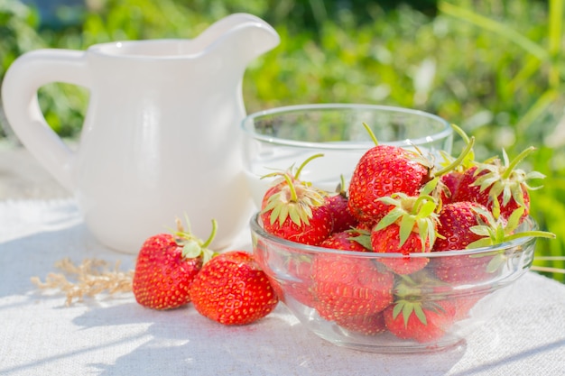 Een kom aardbeien, een beker met melk en een kruik op een servet op een houten tafel