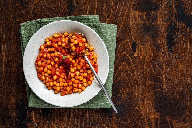 Een kom gestoofde cheak-pea met hete tomatensaus op een servet, op houten achtergrond, overhead geschoten met kopieerruimte.