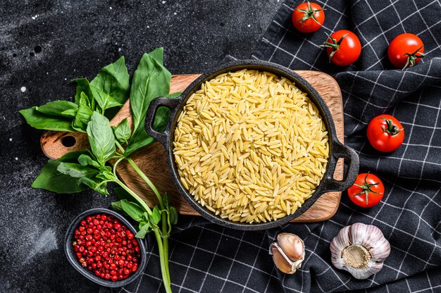 Een kom ongekookte Orzo pasta met basilicumblaadjes, tomaten en knoflook. Zwarte achtergrond. Bovenaanzicht.