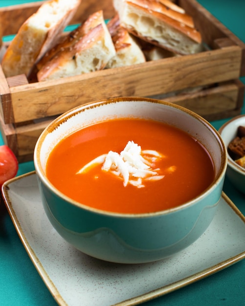 Een kom tomatensoep met brood op de achtergrond.