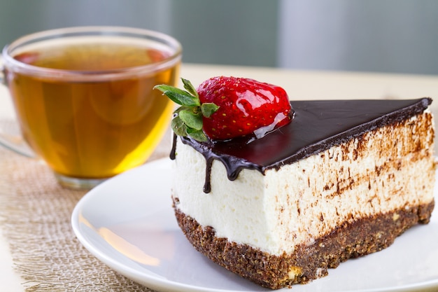 Een kop warme thee en een plakje zoete cake met slagroom, verse aardbeien en druipende chocoladeglazuur in een witte plaat. Zoet dessert