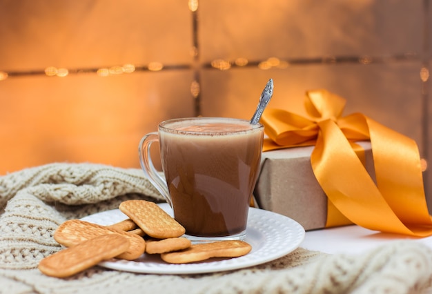 Een kopje cacao en koekjes op een witte plaat op een witte tafel en een geschenk met een gouden lint