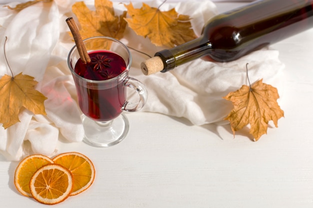 Een kopje glühwein met kruiden, fles, een sjaal, droge bladeren en sinaasappelen op de tafel. Herfststemming, een methode om warm te blijven in de kou, copyspace.