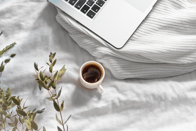Een kopje koffie, een laptop en een groene eucalyptustak tegen de achtergrond van een wit bed en een