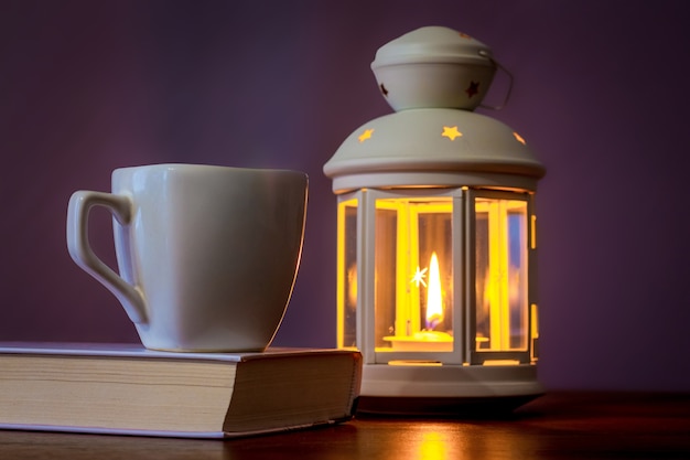 Een kopje koffie op een boek bij een lantaarn met een kaars in de avond