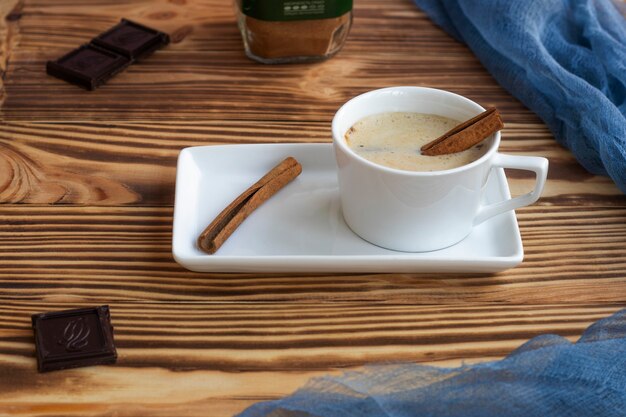 Een kopje latte met schuim staat op een vierkante schotel op een houten tafel Kaneelstokjes donkere chocolade en koffiestukjes