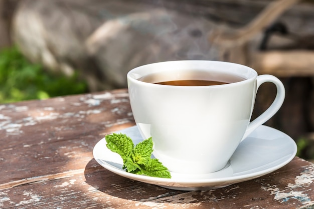 Een kopje thee met een groen blad op oude houten planken en een achtergrond van natuur en platteland.