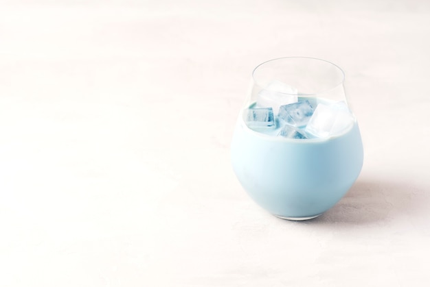 Een koude blauwe matcha latte staat in een glas met ijsblokjes op een grijze tafel.