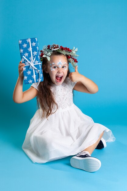 Een lachend meisje in een witte kerstjurk, met een krans en gympen, houdt een geschenkdoos vast