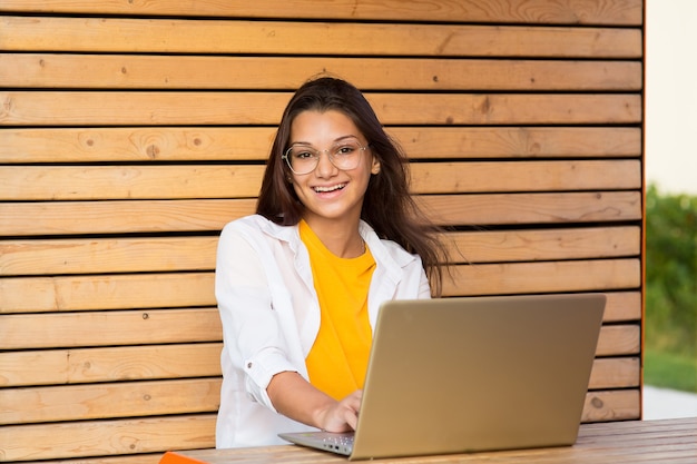 Een lachend meisje met een bril zit op een bankje met een laptop en kijkt in het frame