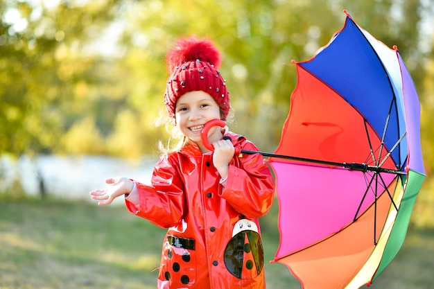 Een lachend meisje met een rode hoed en een rood jasje loopt met een regenboogparaplu in de natuur