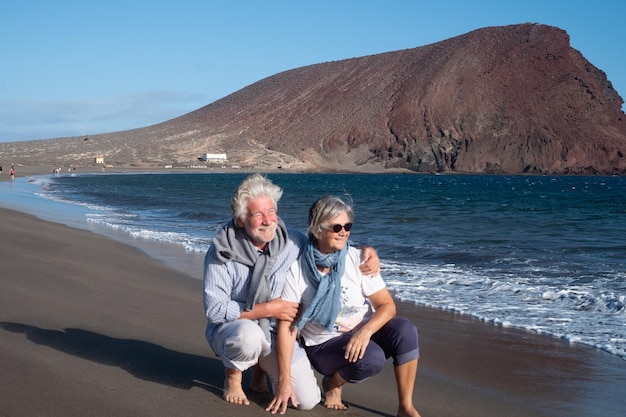 Een lachend senior koppel met grijs haar dat geniet van de zee op een winderige dag. Zeegezicht met een rode berg op de achtergrond