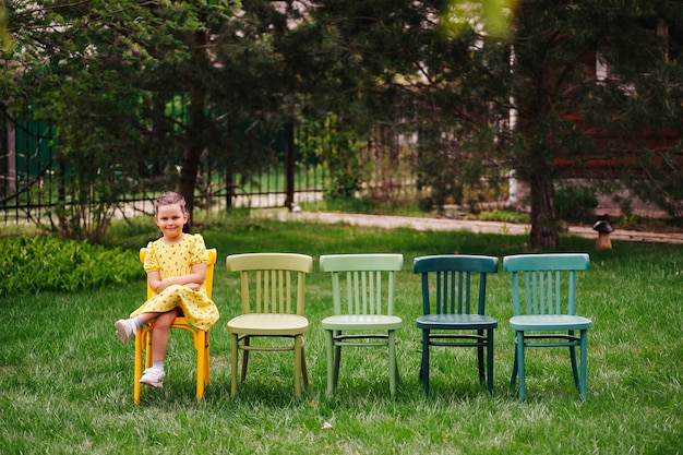 Een lachend vrolijk meisje in een gele jurk zit op Weense veelkleurige geschilderde stoelen te wachten op ...