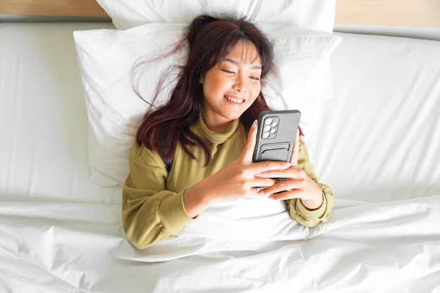 Een lachende jonge Aziatische vrouw lacht terwijl ze haar telefoon vasthoudt en op bed ligt