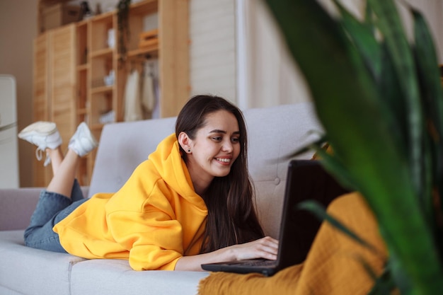 Een lachende jonge brunette vrouw met lang haar in een gele sweater, spijkerbroek en witte sneakers