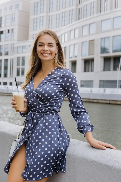 Een lachende jonge vrouw drinkt koffie tegen de achtergrond van moderne stedelijke gebouwen Zomerse stedelijke sfeer